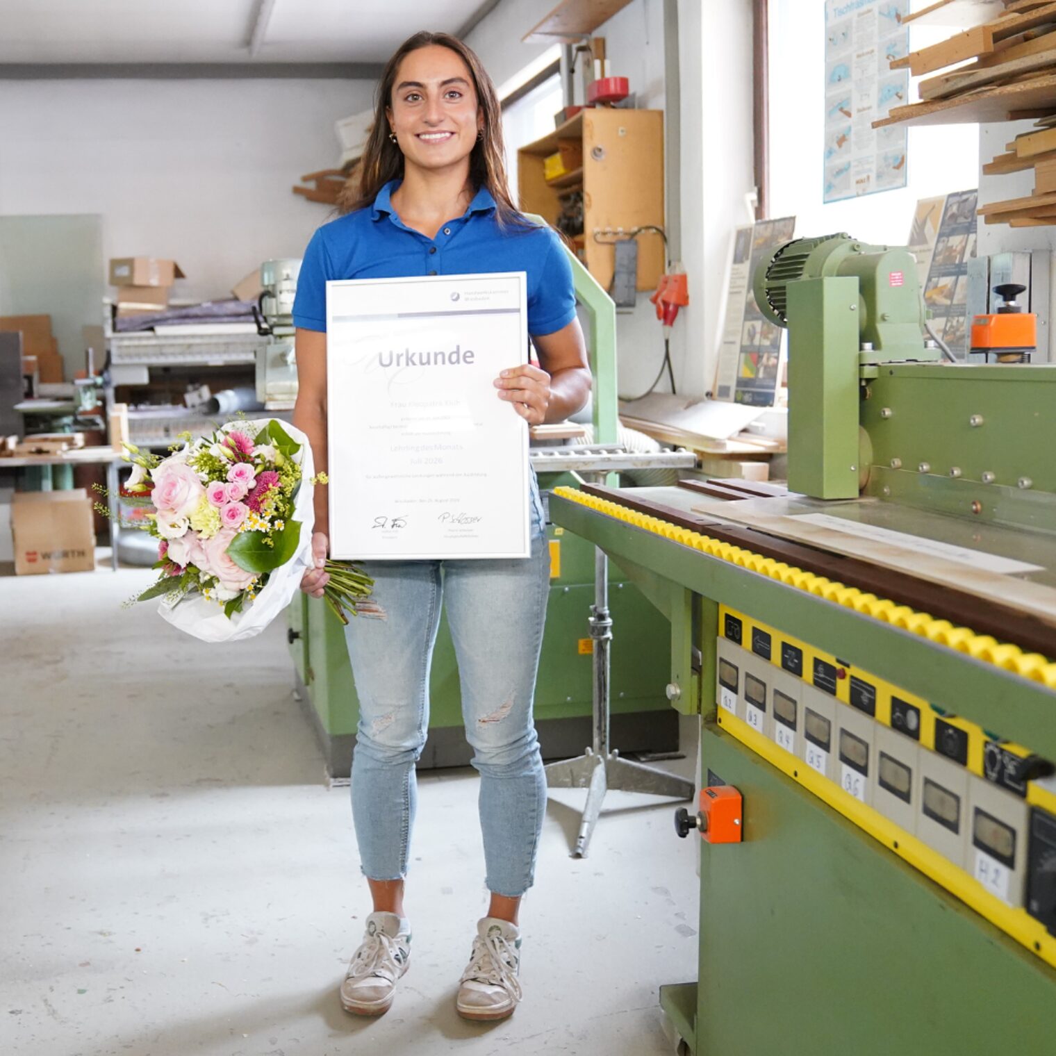 Kleopatra Klüh mit ihrer Urkunde Lehrling des Monats Juli 2026 und einem Blumenstrauß in der Werkstatt stehend.
