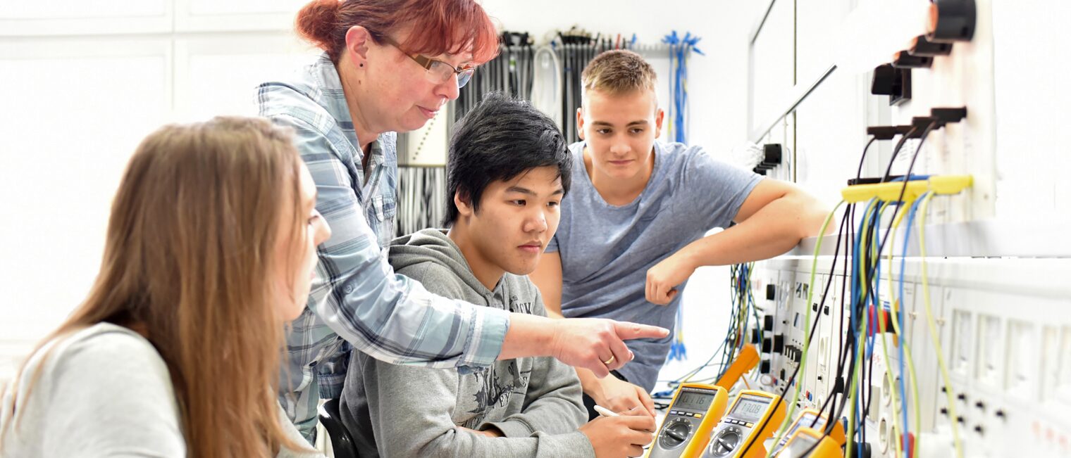 Ausbildungssituation mit Ausbilderin und Azubis in einer Ausbildungswerkstatt für Elektrotechnik